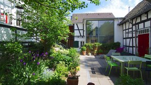 A courtyard with a glass table and chairs, surrounded by potted plants and a large window. - Idyllic, completely renovated half-timbered house in a listed courtyard (Neunkirchen-Seelscheid)