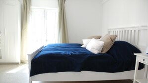 A white bed with a blue bedspread and beige pillows in a room with white walls and a door.