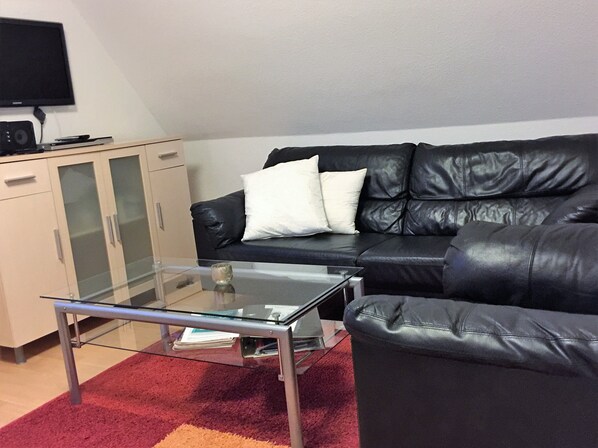 A living room with a glass coffee table, a black leather sofa with white cushions, a beige cabinet, and a flat-screen TV mounted on the wall.