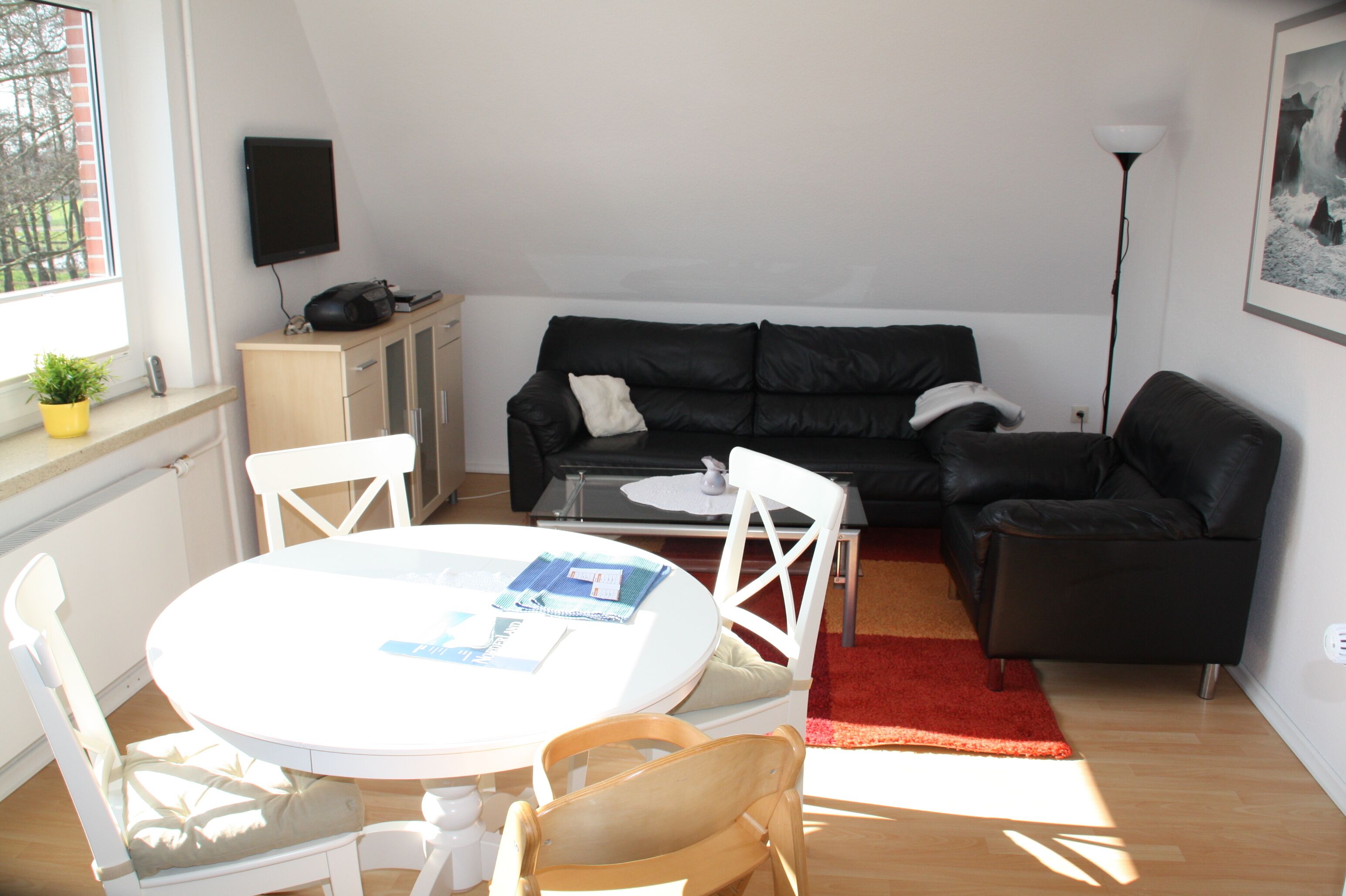 Un salon avec un canapé en cuir noir, une table à manger blanche avec des chaises, une petite table blanche et un tapis rouge.