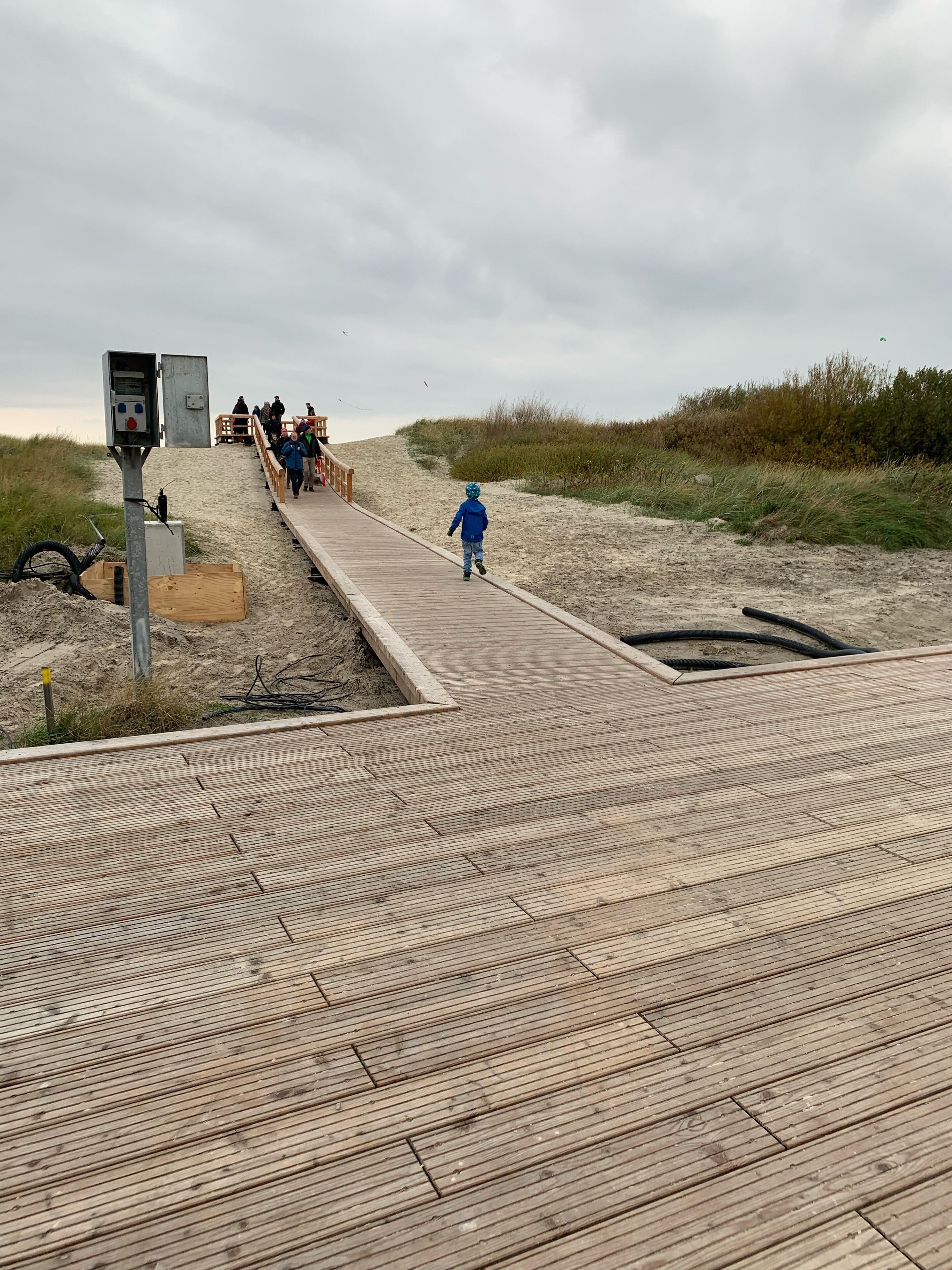 Ein hölzerner Steg führt zu einem Sandstrand, im Hintergrund sind ein Schaltkasten und ein Holzgebäude zu sehen.