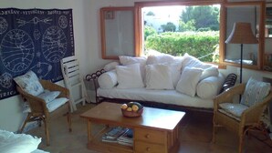 Una sala de estar acogedora con un sofá blanco, dos sillones de mimbre, una mesa de centro de madera y un ventanal con vistas a zonas verdes.