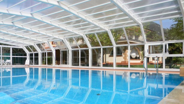 Une piscine dotée d’un toit en verre, entourée de chaises longues et offrant une vue sur un bâtiment avec un escalier.