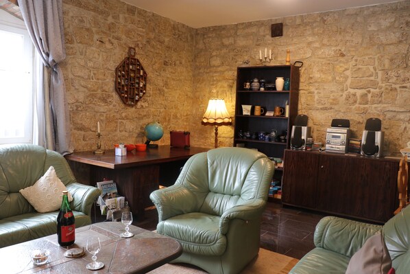 A cozy living room with a stone wall, leather armchairs, a wooden coffee table, and a bookshelf with various items.