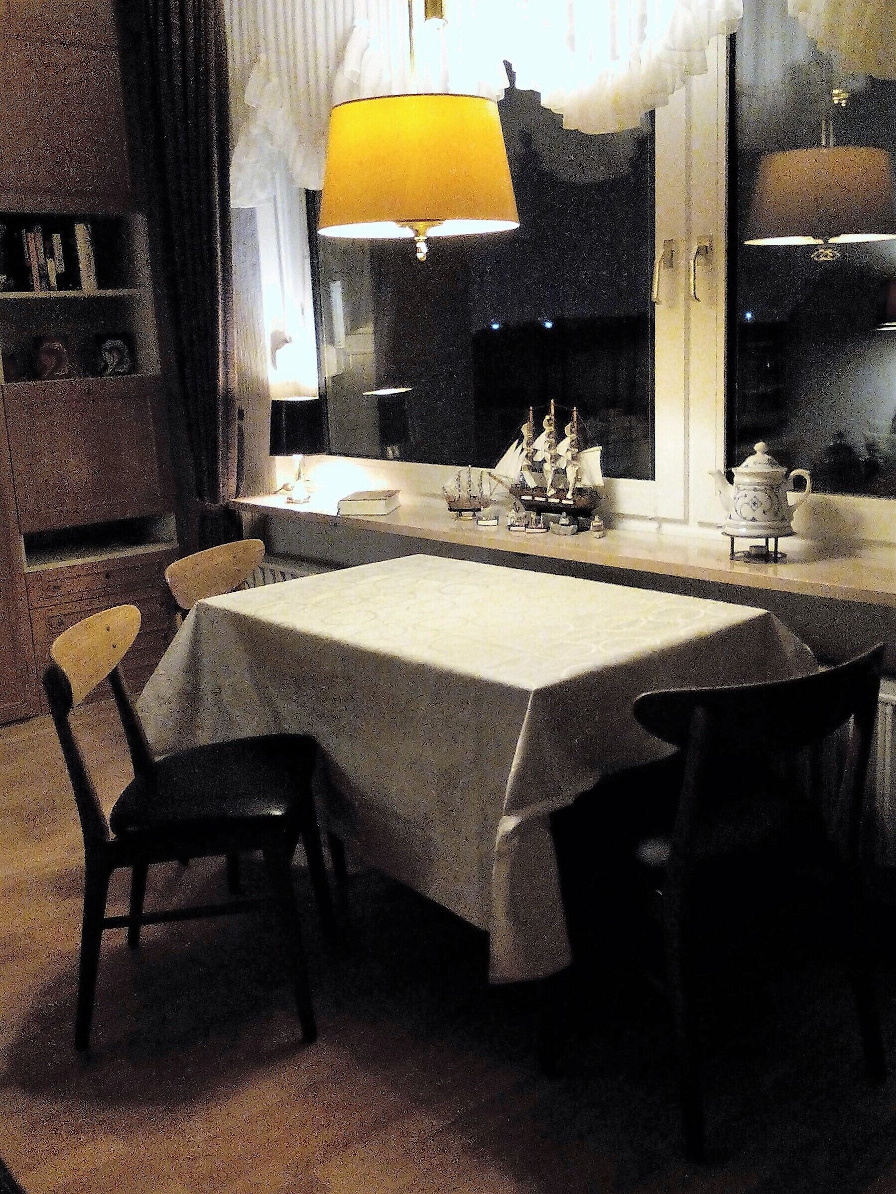 A dining area with a table set for two, chairs, a lamp, and a window with a view.