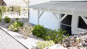 A white building with a covered porch, gravel pathway, and various shrubs and plants.