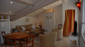 Ein gemütliches Wohnzimmer mit einem Holzesstisch, einer Couch und einem Sessel mit Bezug. Ein Fernseher steht auf einem kleinen Tisch und ein Schrank mit einem herzförmigen Spiegel ist ebenfalls zu sehen.