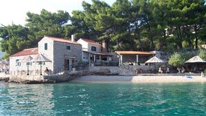 A stone building with a red roof, a balcony, and a beach area with umbrellas. - The apartment is located on the first floor of our house, 50 meters from the sea (Bol)
