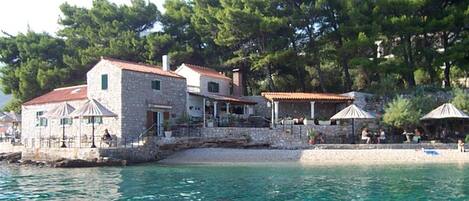 A stone building with a red roof, a balcony, and a beach area with umbrellas.