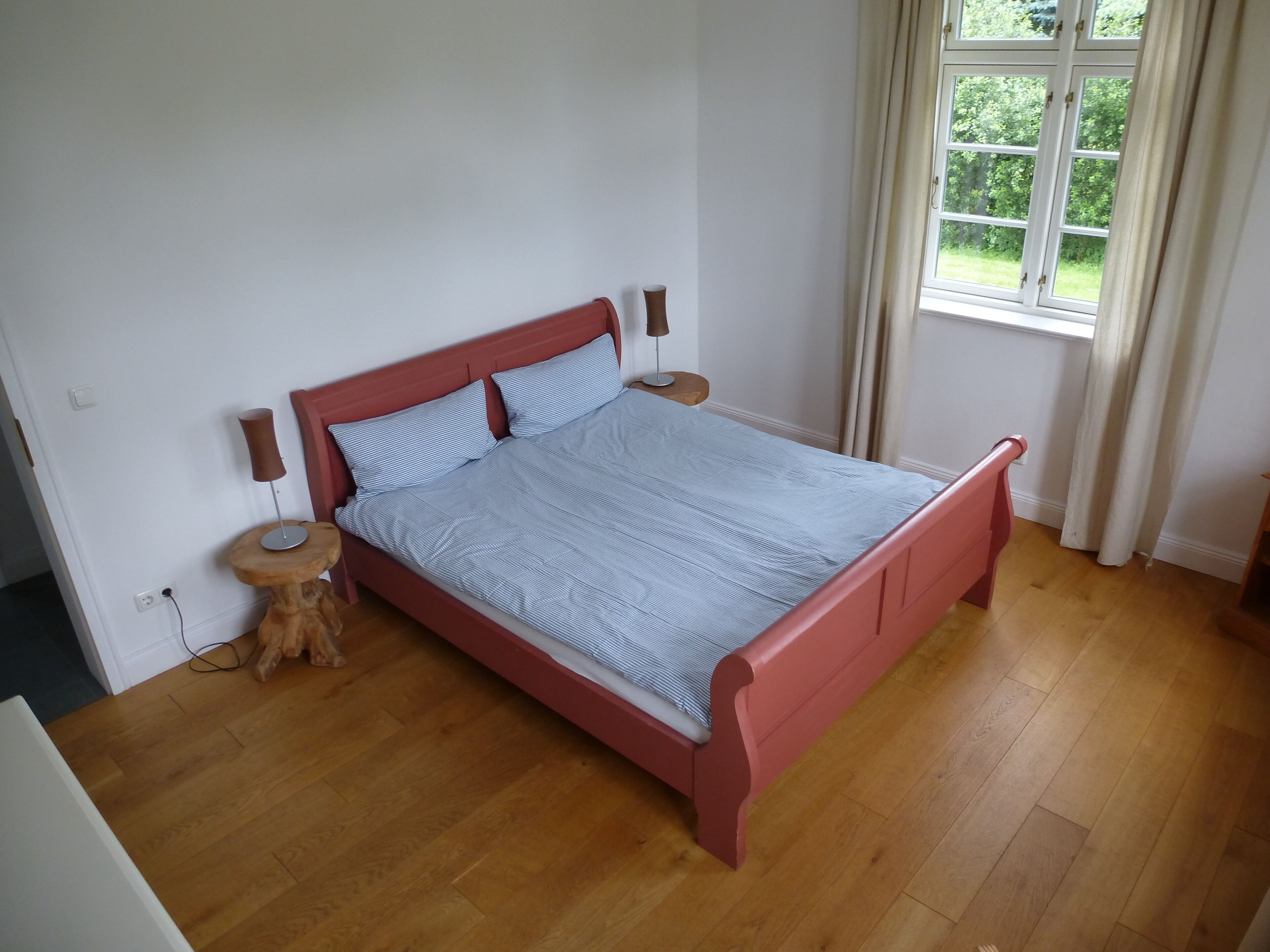 A bedroom with a wooden floor, a bed with a red frame, two bedside tables, and a window with curtains.