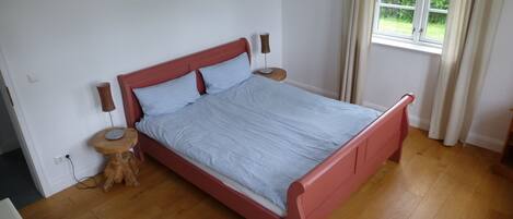 A bedroom with a wooden floor, a bed with a red frame, two bedside tables, and a window with curtains.