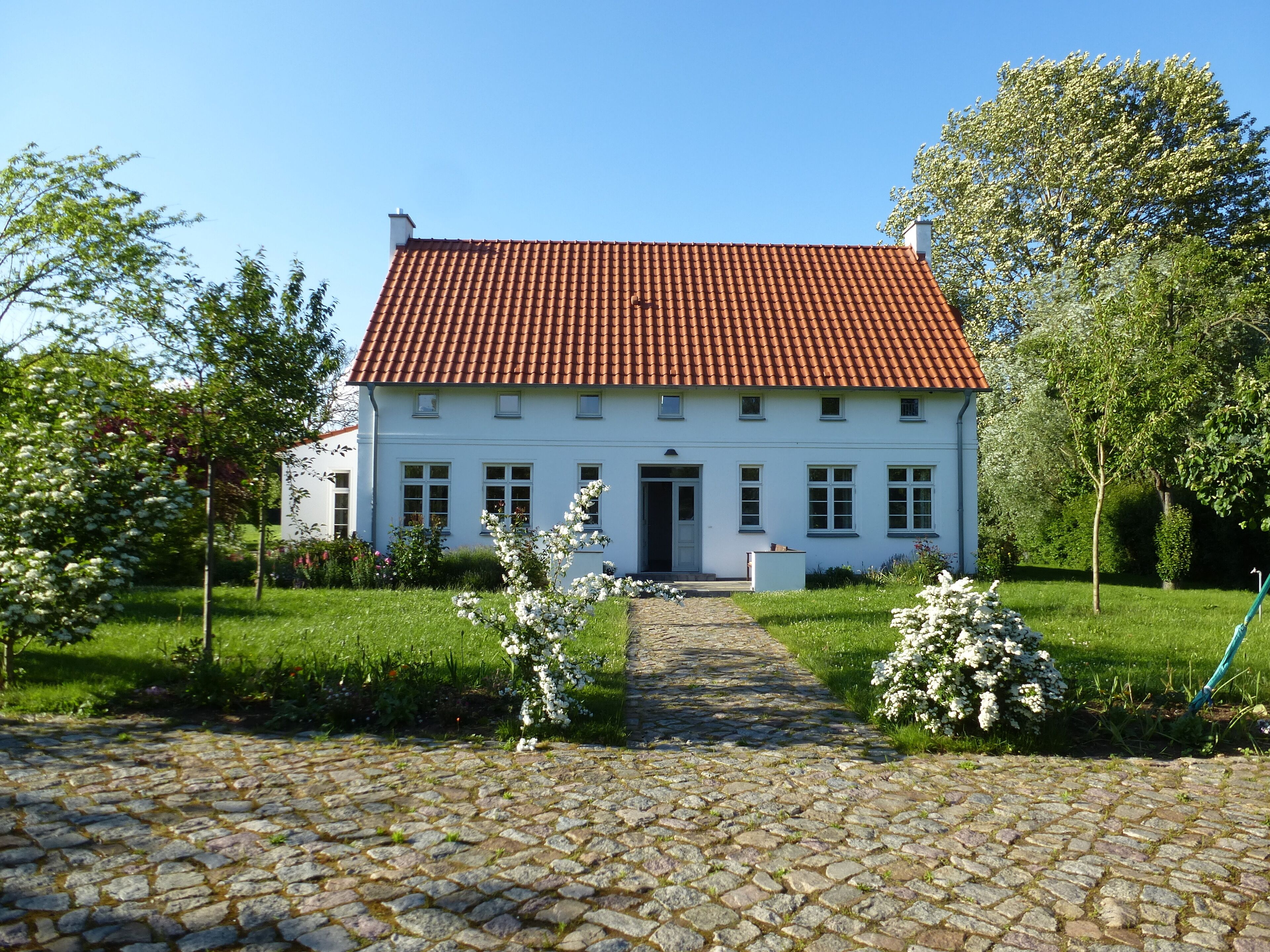 A white building with a red-tiled roof, a cobblestone driveway, and a garden with white flowering plants.
