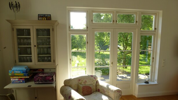 Una habitación con piso de madera, un sillón floral, un pequeño reposapiés, un escritorio con alacena y un amplio ventanal con vistas a follaje.