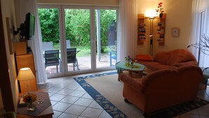 Un salon comprenant un canapé, une petite table avec une lampe et une vue sur un jardin, accessible par une porte-fenêtre coulissante.
