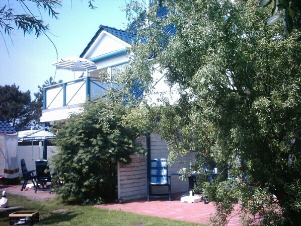 A two-story house with a covered patio area, a large tree in front, and a blue barrel. - Comfortable, ground-floor vacation apartment with terrace and garden to make you feel at home. (24217 Schönberger Strand)