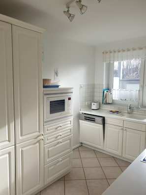A kitchen with white cabinetry, a microwave, a dishwasher, and a window with a view of trees. - Comfortable, ground-floor vacation apartment with terrace and garden to make you feel at home. (24217 Schönberger Strand)
