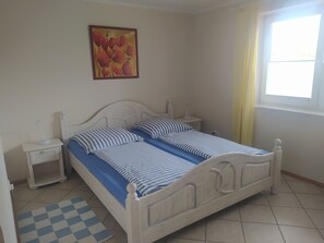 Un dormitorio con cama doble, mesita de noche, lámpara y alfombra a cuadros, además de una ventana con cortinas.