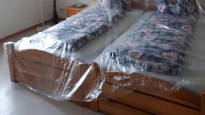 Une pièce avec un lit en bois recouvert de plastique, un tableau accroché au mur et une fenêtre.