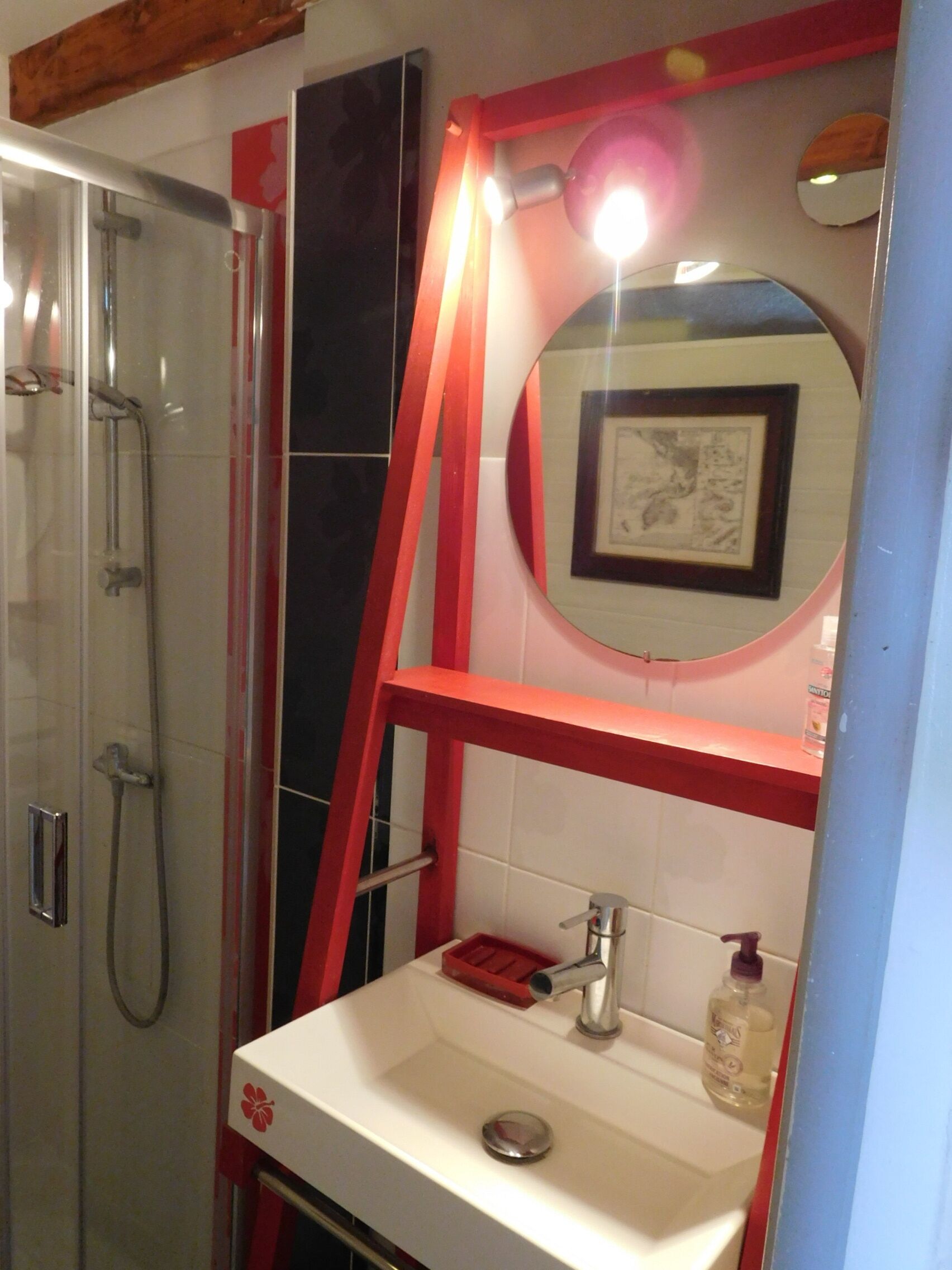 A bathroom with a glass shower enclosure, a white sink with a red soap dispenser, and a framed picture on the wall.