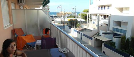 Ein Balkon mit Blick auf das Meer und Gebäude. Es gibt Stühle und einen Tisch mit einer Plastiktüte darauf.