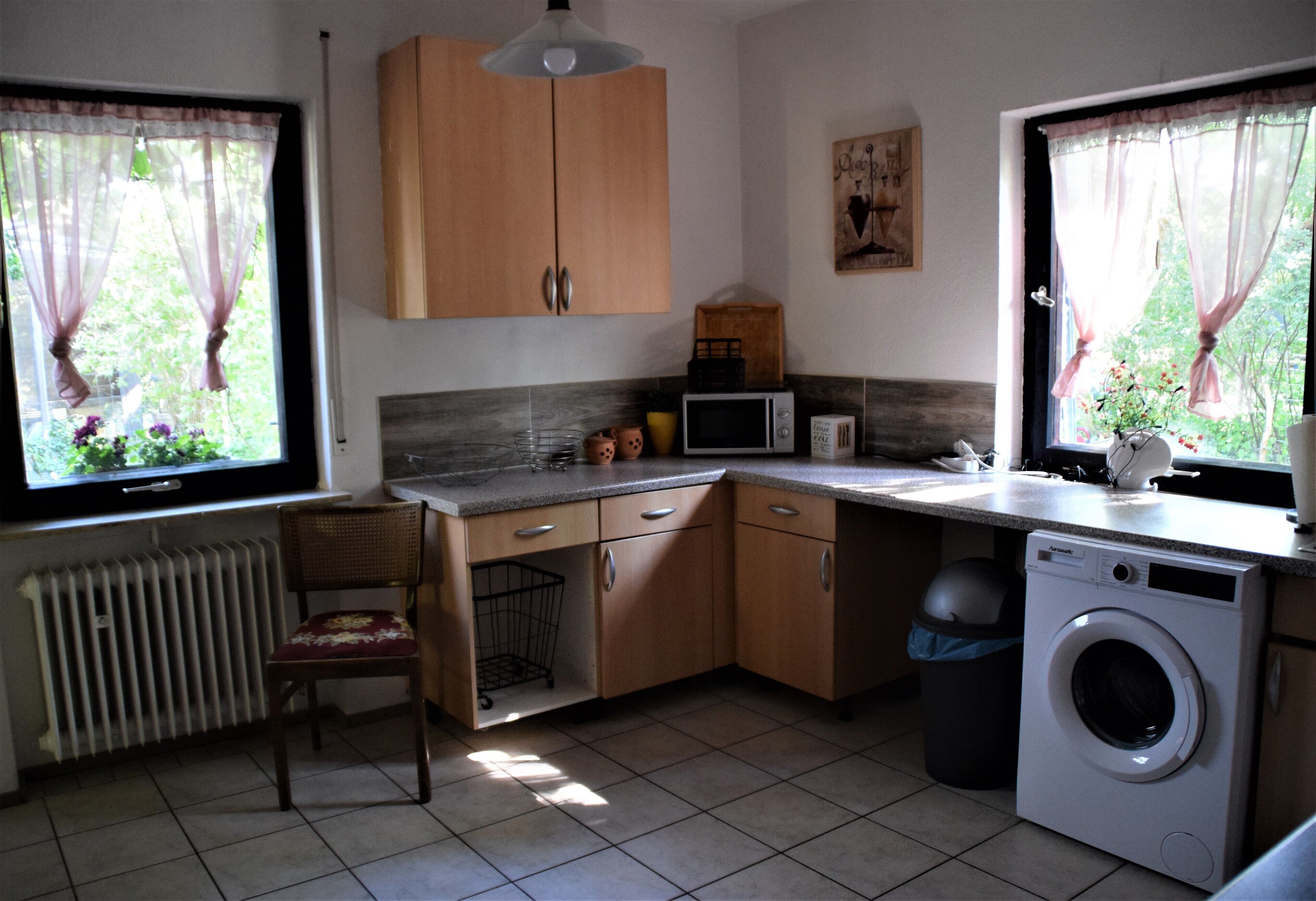 Cocina compacta con armarios de madera, microondas, lavadora y una silla.