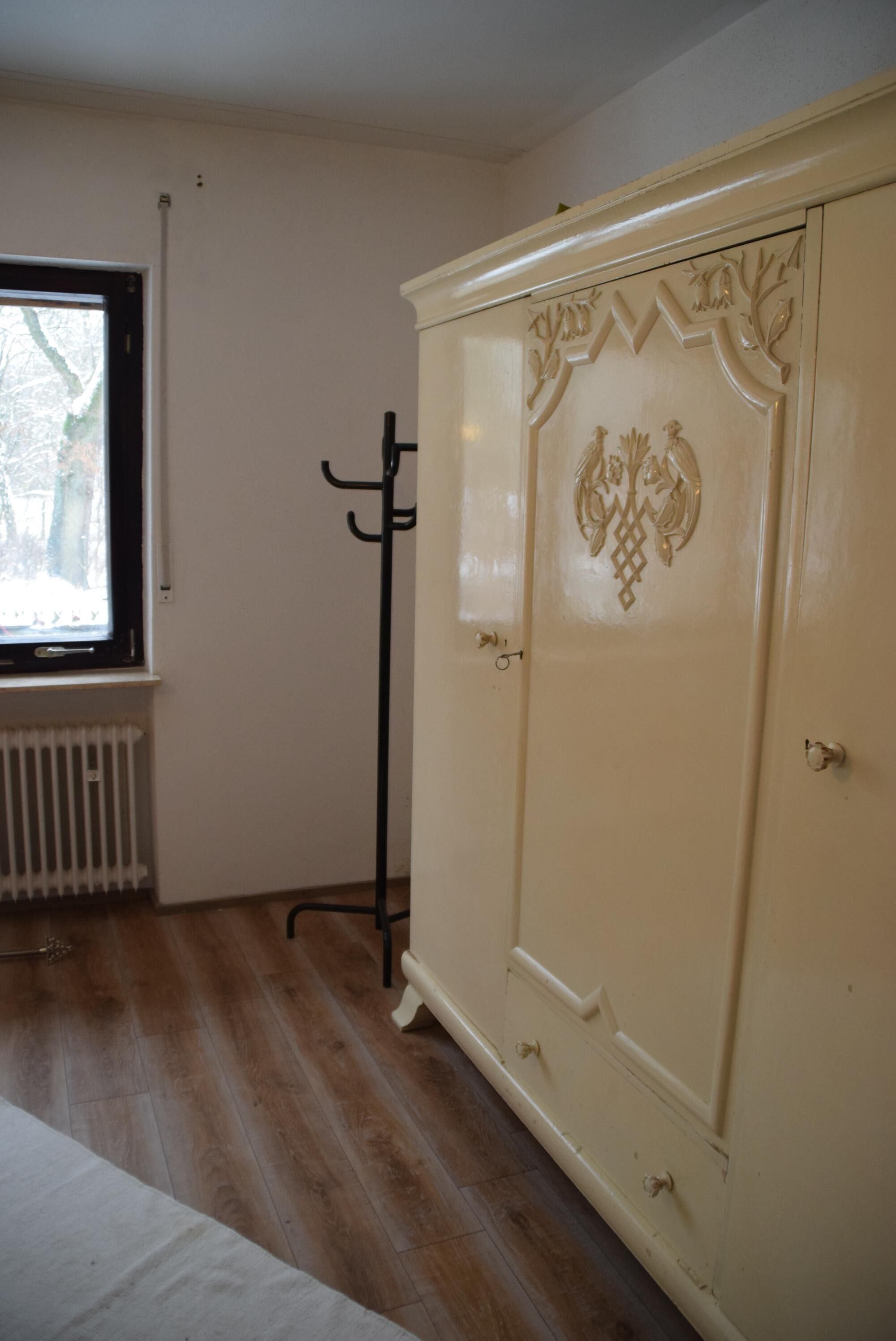 Quarto com piso de madeira, radiador, janela com vista para árvores e guarda-roupa bege com detalhes entalhados.