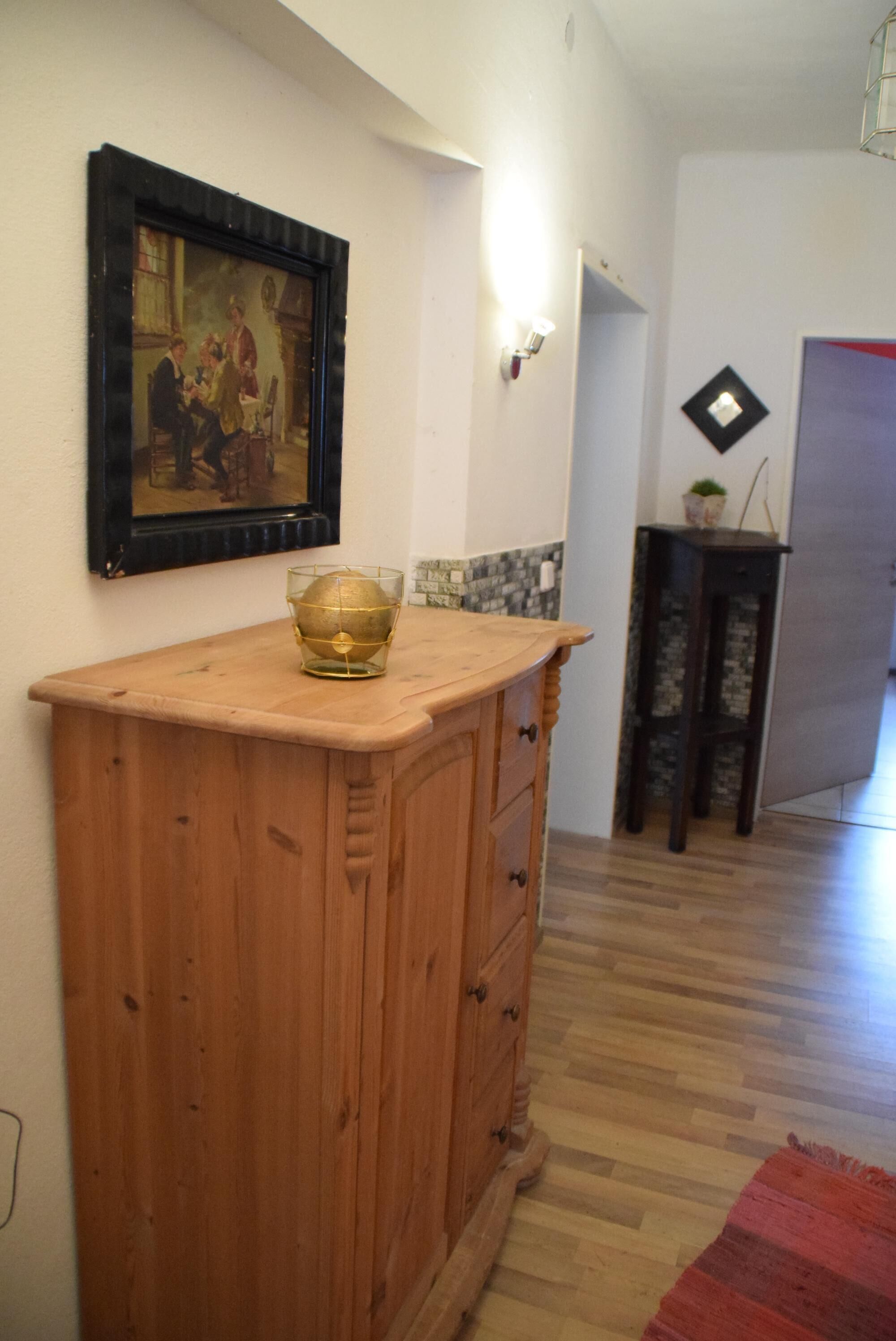 Een houten kast met lades, een lijst met een schilderij aan de muur en een klein tafeltje met een plant.