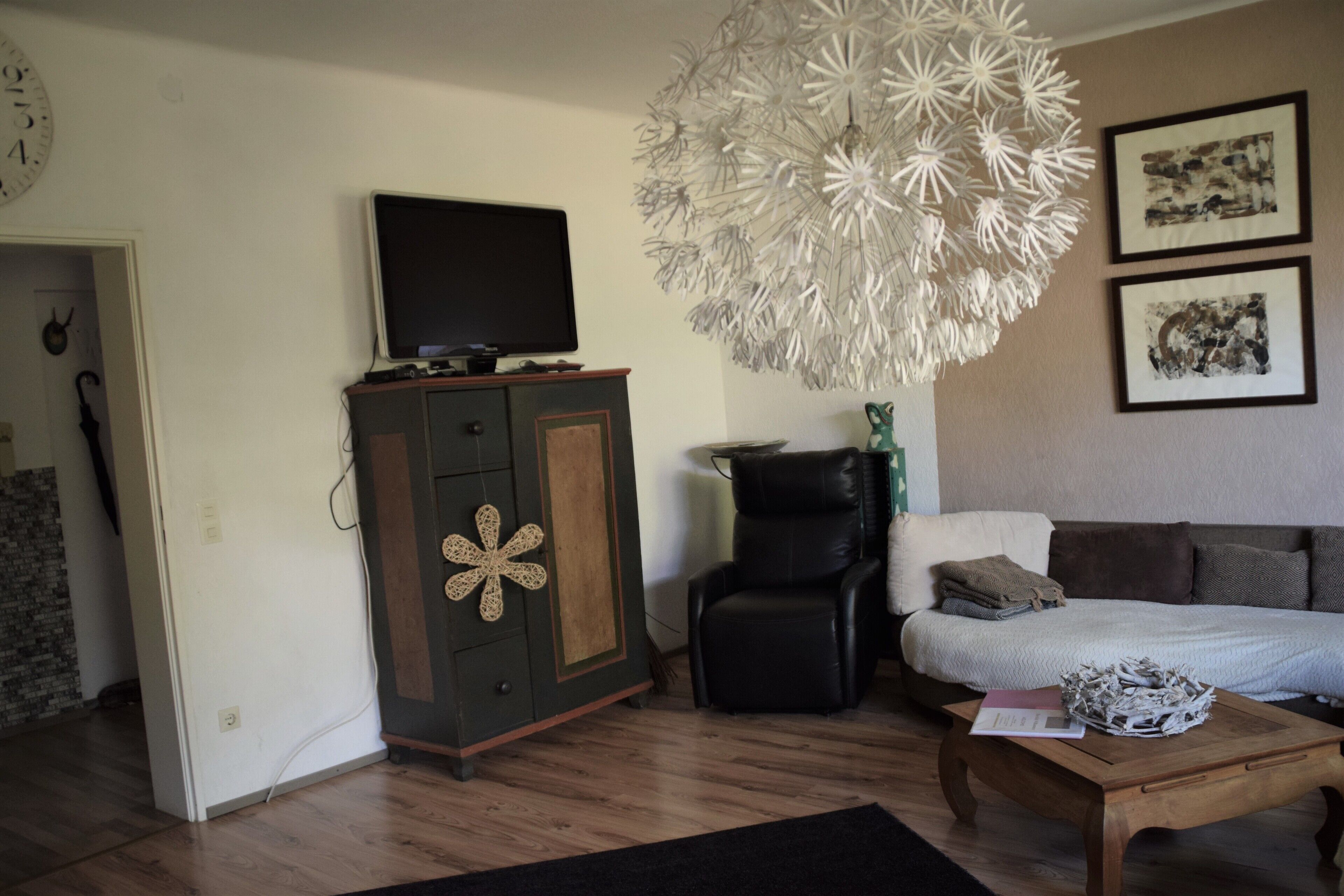 A living room with a television on a cabinet, a black leather chair, a white sofa, a wooden coffee table, and a decorative chandelier.