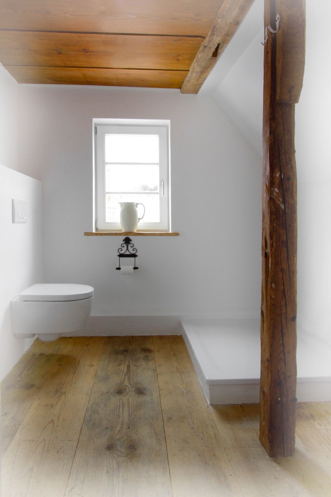Une petite salle de bain blanche, avec un plancher en bois, des toilettes blanches, une fenêtre et un pichet blanc posé sur une étagère.