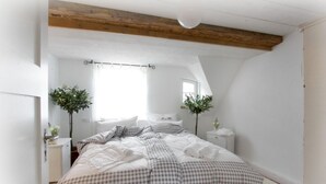 A bedroom with a bed, white walls, a wooden ceiling beam, a window with curtains, and two bedside tables with plants. - Deluxe farmhouse, smokehouse kitchen, tiled stove, bathroom in stable, Whirlpool (Bubsheim)