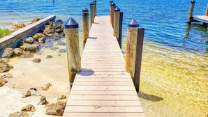 Eine Holzstege führt ins klare, blaue Wasser zu einem sandigen Strand auf der linken Seite, während sich in der Ferne die Skyline einer Stadt abzeichnet.