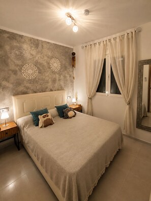 Une chambre à coucher avec un grand lit, une table de chevet avec une lampe, une fenêtre avec des rideaux et un mur décoré d’assiettes.