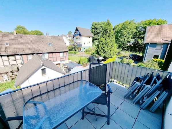 Un balcon avec une table et des chaises en verre, une clôture en métal et une vue sur les maisons et les arbres des environs.