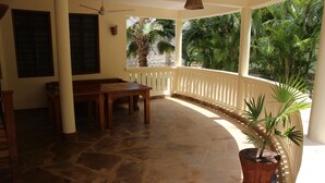 A covered patio with a wooden table and chairs, a potted plant, and a view of palm trees.