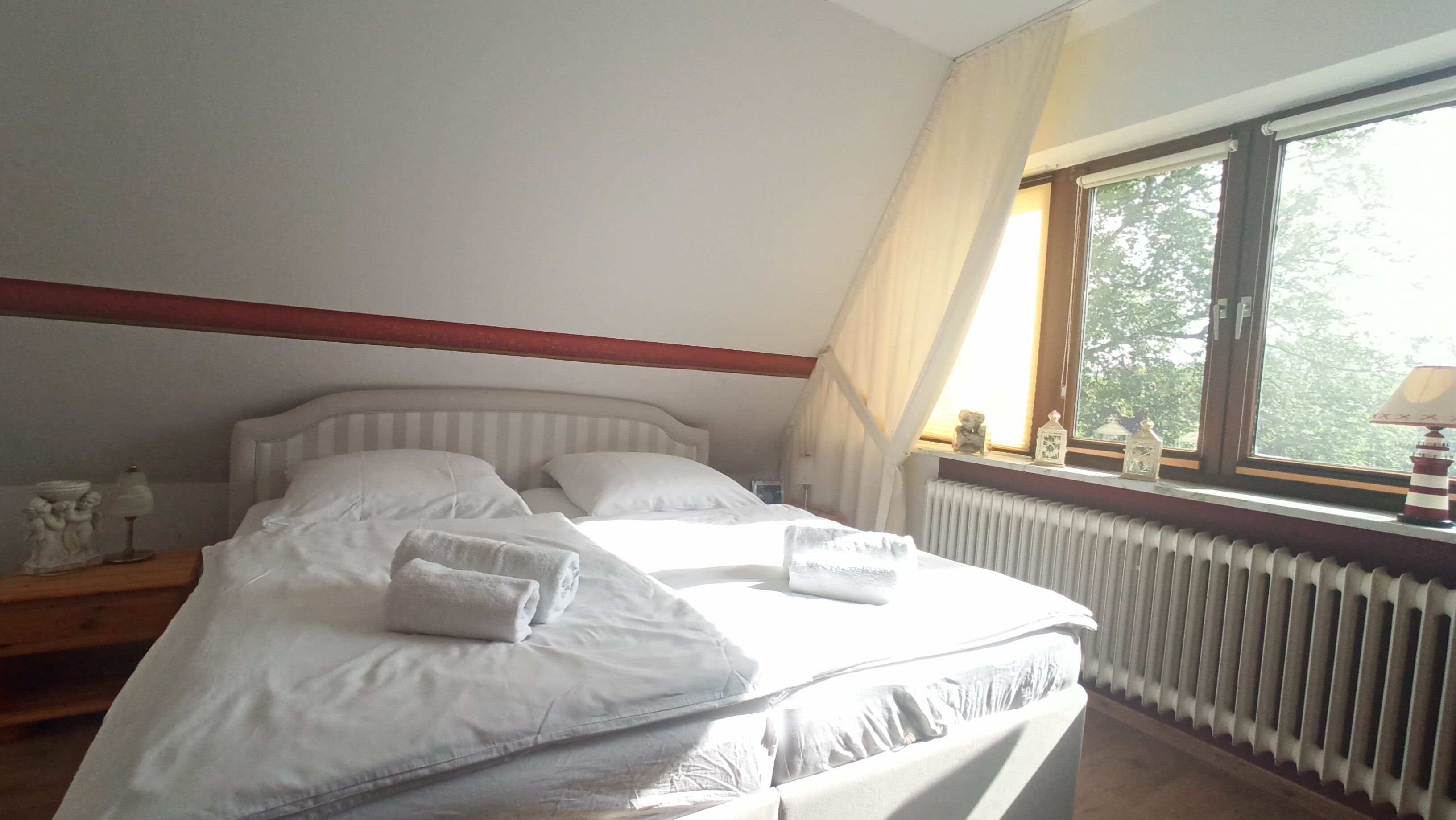A neatly made bed with white linens and towels, a wooden headboard, a window with curtains, and a radiator below.