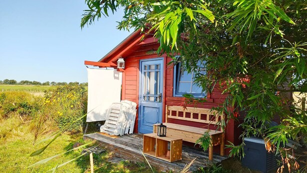 Une petite maison en bois rouge, avec une porte et une fenêtre bleues, un banc en bois et un tissu blanc accroché à la porte.