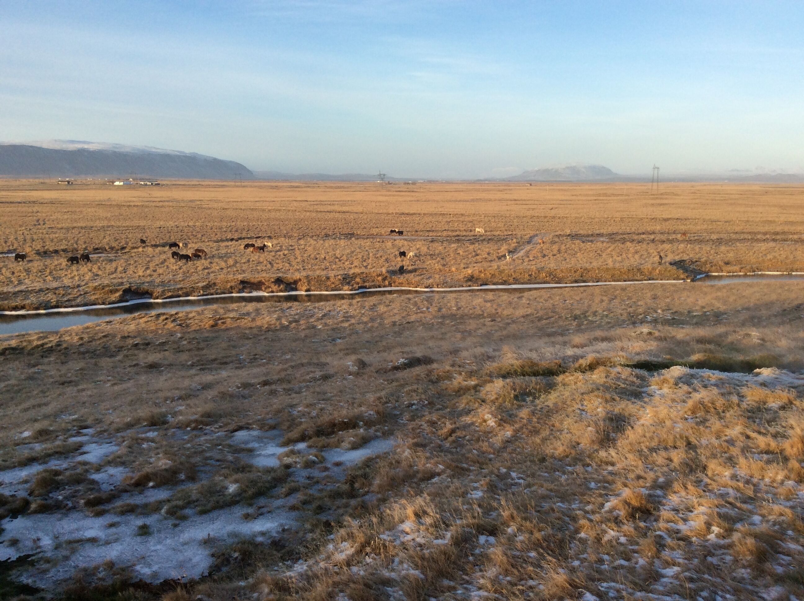 A vast, open landscape with a river running through it, surrounded by dry grass and snow patches.
