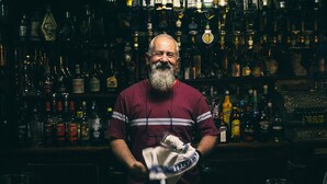 Ein Mann mit kariertem Hemd hält ein Handtuch in einer Bar, in der Regale mit Spirituosenflaschen stehen.
