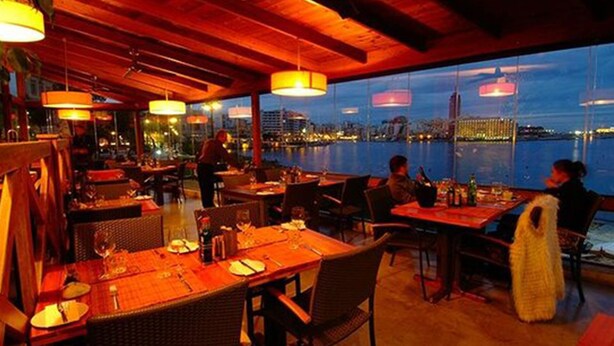 Ein Restaurant im Freien mit Tischen und Stühlen, Blick auf das Wasser und hängende Lichter.