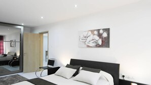 Ein modernes Schlafzimmer mit einem großen Bett, einem Schreibtisch und einem Stuhl. An der Wand hängt ein Gemälde und eine Tür führt in einen weiteren Raum.