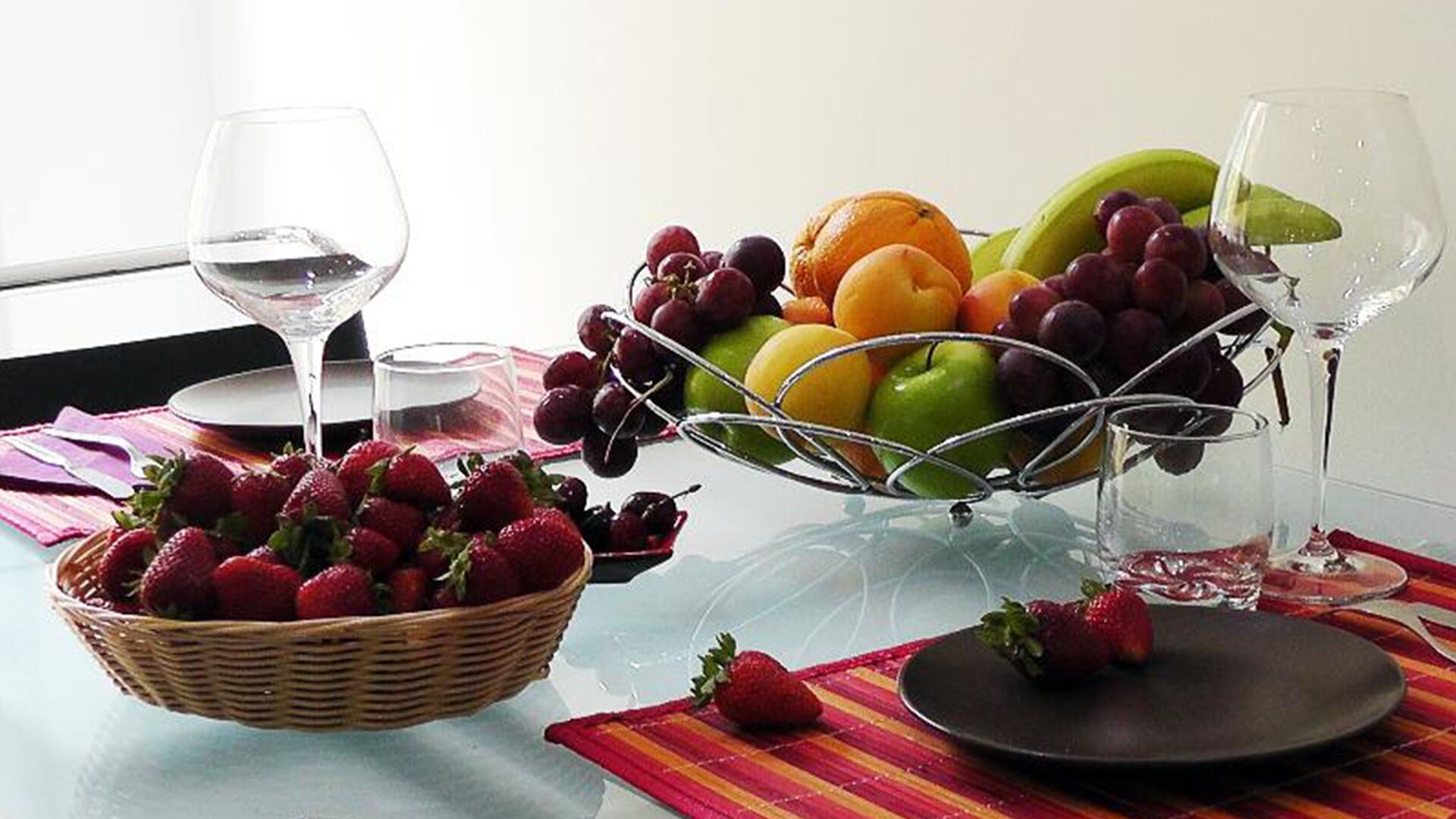 A table with a fruit basket, two wine glasses, and a plate with a strawberry.
