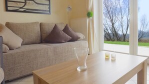 A living room with a sofa, a coffee table, a lamp, and a view of trees through a window. - Newly furnished apartment with a large terrace (Ostseebad Boltenhagen)