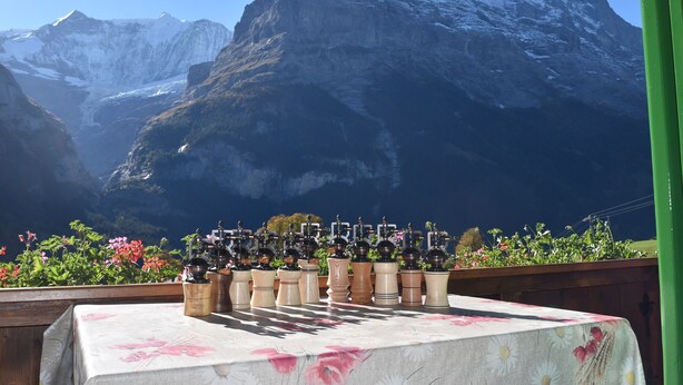 A chessboard set on a table with a floral tablecloth, surrounded by a wooden fence and colorful flowers, with a mountain backdrop.