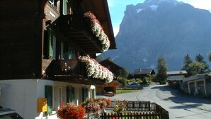 A traditional chalet with a balcony adorned with flowers, a mountain backdrop, and a clear sky.