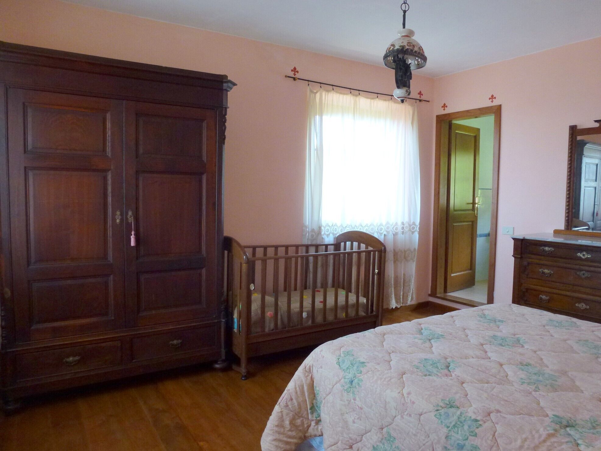 A room with a large wooden wardrobe, a crib, a bed with a floral bedspread, a mirror, and a door leading to another room.