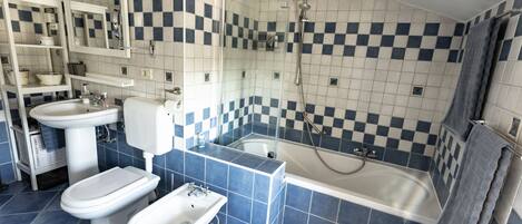 Une salle de bain avec un carrelage à motifs à carreaux, une baignoire, deux toilettes, un lavabo et un miroir.