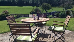 A round wooden table with chairs, a wine bottle, and a glass, set on a gravel patio with a view of a green field and trees. - Welcome to the Allgäu family Berger (Sulzberg)