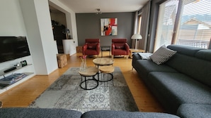 Een moderne woonkamer met een grijze bank, twee rode fauteuils, een salontafel en een televisie op een plank.