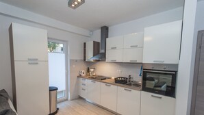 A modern kitchen with white cabinets, a stainless steel range hood, a built-in oven, and a window with blinds.