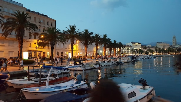 A marina with boats docked along a waterfront, surrounded by buildings and palm trees.